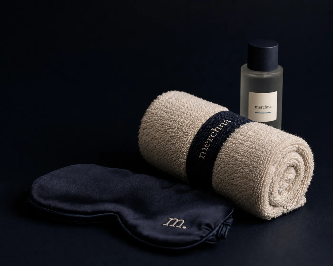 A rolled Merchna towel with a branded band, an amenity bottle and a silk eye mask